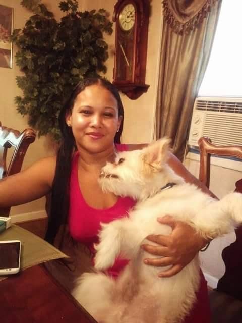 Teola Blake in a red top, seated at a dining table holding her small white dog.